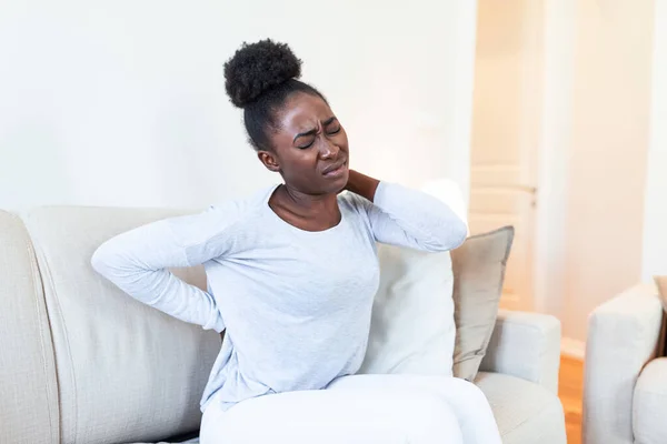 Young Black woman suffering from backache at home. Portrait of a young brunette girl sitting on the couch at home with a headache and back pain. Beautiful woman Having Spinal Or Kidney Pain
