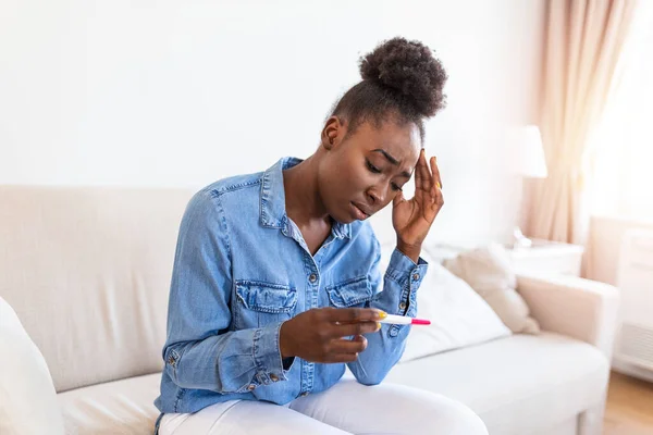 Sad, worried african american woman checking her recent pregnancy test, sitting on couch at home. Maternity, child birth and family problems concept. unwanted pregnancy