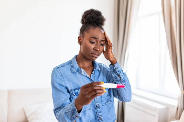 Sad, worried african american woman checking her recent pregnancy test, sitting on couch at home. Maternity, child birth and family problems concept. unwanted pregnancy