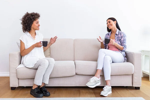 Two female friends in social distancing sitting on sofa in coronavirus pandemic time. Best friends having coffee together while separated by social distancing on sofa at home