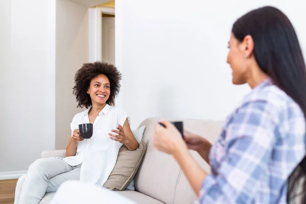 Two female friends in social distancing sitting on sofa in coronavirus pandemic time. Best friends having coffee together while separated by social distancing on sofa at home