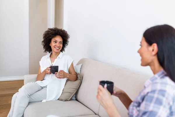 Two female friends in social distancing sitting on sofa. Best friends having coffee together while separated by social distancing on sofa at home