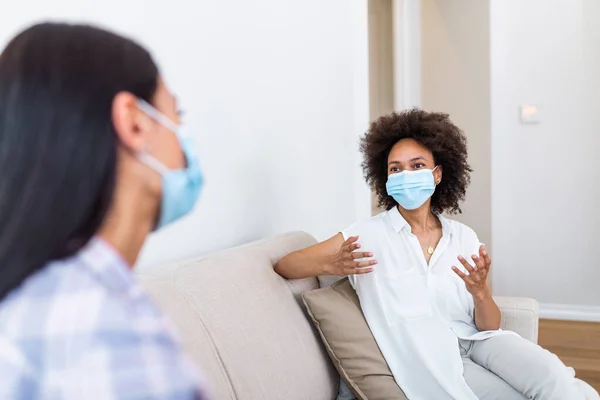 Two female best friends sitting in social distance wearing face mask and talking on the sofa, preventing covid 19 coronavirus infection spread.