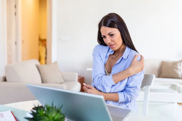 Portrait of young stressed woman sitting at home office desk in front of laptop, touching aching shoulder with pained expression, suffering from shoulder after working on laptop