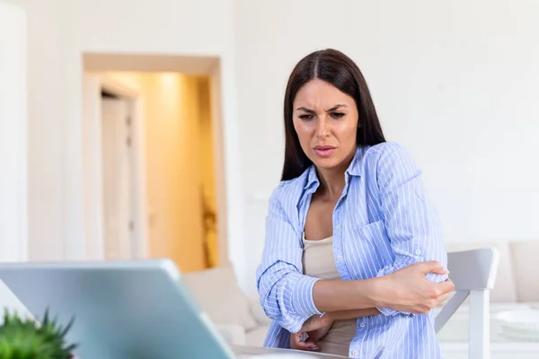 Portrait of young stressed woman sitting at home office desk in front of laptop, touching aching elbow with pained expression, suffering from elbow after working on laptop