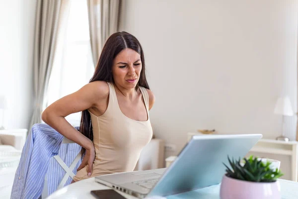 Portrait of young stressed woman sitting at home office desk in front of laptop, touching aching back with pained expression, suffering from backache after working on laptop