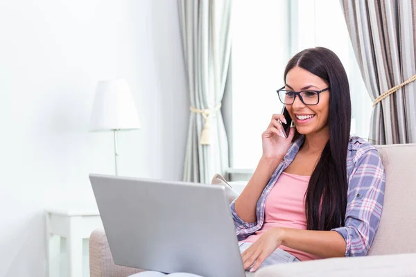 Happy woman sitting on sofa with laptop and talking on phone at home. Young successful businesswoman working from home while talking at phone. College student studying on laptop and using phone.