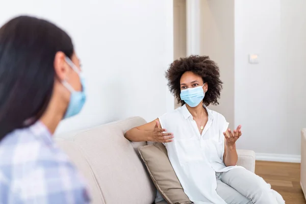 Two female best friends sitting in social distance wearing face mask and talking on the sofa, preventing covid 19 coronavirus infection spread.