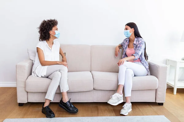 Two female friends in social distancing sitting on sofa in coronavirus pandemic time. Best friends having coffee together while separated by social distancing on sofa at home
