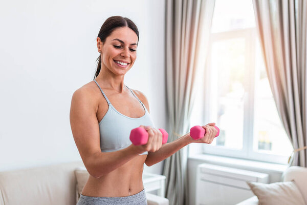 Woman exercising with dumbbells at home. Sporty beautiful woman exercising at home to stay fit. Young woman exercising at home in a living room. Fitness, workout, healthy living and diet concept.