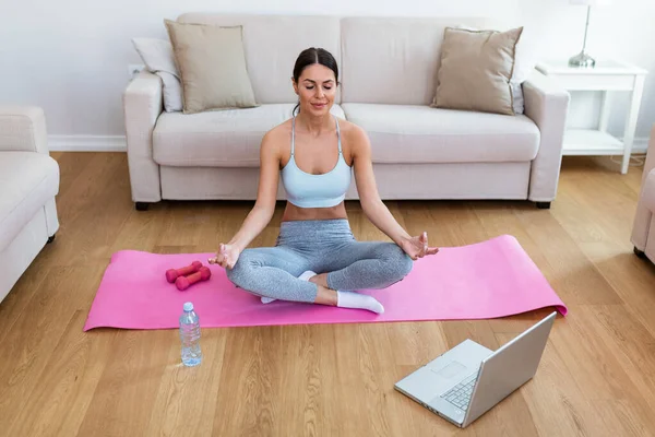 Self learning. Woman using laptop to training yoga online in living room. Girl does yoga training exercises on a mat in her living room. She follows an online exercise course video on her laptop.