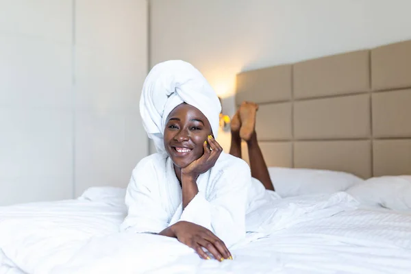 Beautiful young African woman in bathrobe resting on bed after shower at home. Young black woman wearing a towel and a white bathrobe on the bed at home