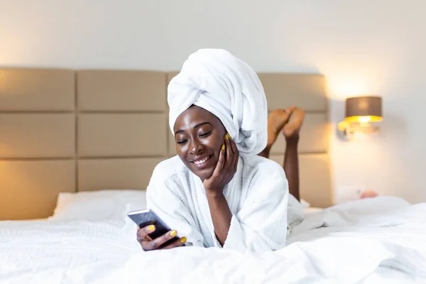Attractive young black girl in bathrobe and with a towel on her head is holding mobile phone,texting and smiling while sitting on a bed. Young African woman on bed