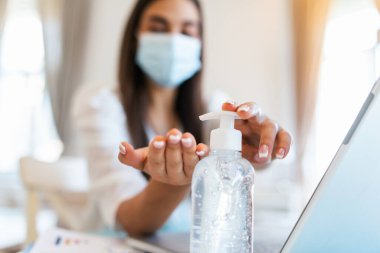 Closeup of woman hand using sanitizer gel to disinfect hands with alcohol gel and face mask over a work desk in office. Preventive measures during the period of pandemic and social exclusion.