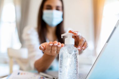 Young woman with face mask back at work in office after lockdown, disinfecting hands. Female employee in protective face mask sanitize hands with antibacterial liquid, protect from COVID-19 pandemic