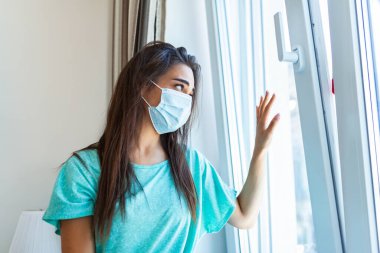 Young woman wearing protective face mask looking outside window with sadness in her eyes, self isolation due to the global COVID-19 Coronavirus pandemic