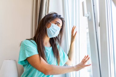Young woman wearing protective face mask looking outside window with sadness in her eyes, self isolation due to the global COVID-19 Coronavirus pandemic