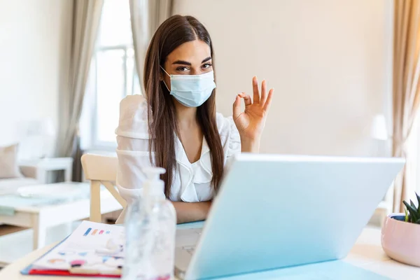 Quarantine, remote job and pandemic concept - woman wearing face protective medical mask for protection from virus disease with laptop computer showing OK sign