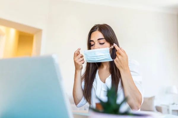 Businesswoman putting on protective mask working on laptop in the office. Focused company employee worker in protective facial mask working on laptop at home keeping covid 19 virus quarantine measures