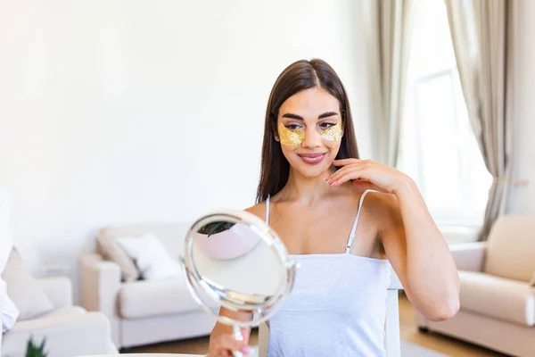 Smiling young woman applying eye hydration mask under her eyes. Female enjoy making face massage, peeling, put on mask under eyes in front of the mirror. Young woman looking her self in the mirror