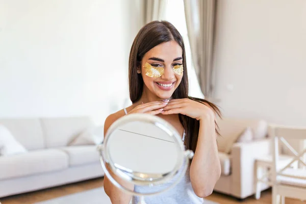 Portrait of pretty, nice young woman with golden eye patches, having bags under eyes in the morning because of less sleep, showing an effect of perfect skin. Looking in the mirror