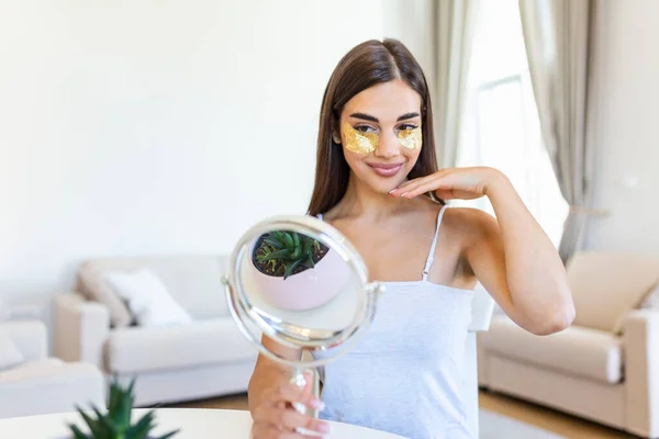 Smiling young woman applying eye hydration mask under her eyes. Female enjoy making face massage, peeling, put on mask under eyes in front of the mirror. Young woman looking her self in the mirror