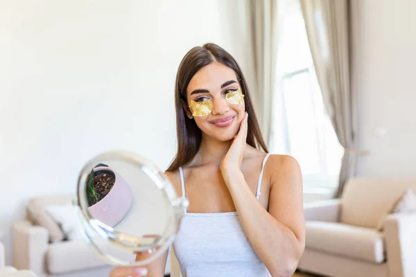 Smiling young woman applying eye hydration mask under her eyes. Female enjoy making face massage, peeling, put on mask under eyes in front of the mirror. Young woman looking her self in the mirror