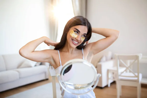 Smiling young woman applying eye hydration mask under her eyes. Female enjoy making face massage, peeling, put on mask under eyes in front of the mirror. Young woman looking her self in the mirror