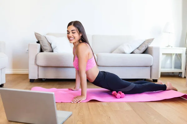 Girl training at home, doing workout and watching videos on laptop, training in living room. Beautiful woman is doing side plank exercise. She is watching video on the internet and repeating the tasks