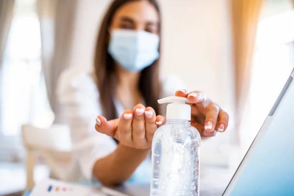 Young woman with face mask back at work in office after lockdown, disinfecting hands. Female employee in protective face mask sanitize hands with antibacterial liquid, protect from COVID-19 pandemic