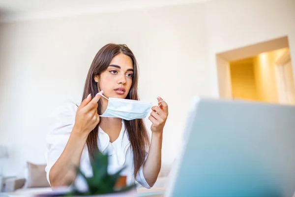 Businesswoman putting on protective mask working on laptop in the office. Focused company employee worker in protective facial mask working on laptop at home keeping covid 19 virus quarantine measures