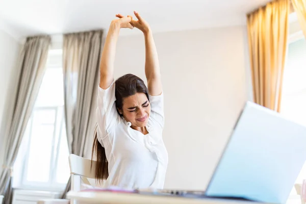 Portrait of young stressed woman sitting at home office desk in front of laptop, streaching her back with pained expression, suffering from backache after working on pc