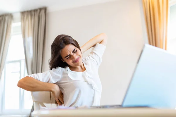 Portrait of young stressed woman sitting at home office desk in front of laptop, touching aching back with pained expression, suffering from back, spine pain after working on pc
