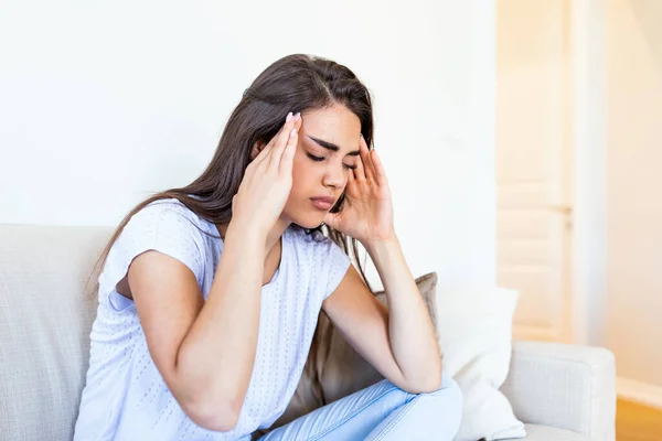 Portrait of an attractive woman sitting on a sofa at home with a headache, feeling pain and with an expression of being unwell. Upset woman sitting on couch feeling strong headache migraine.