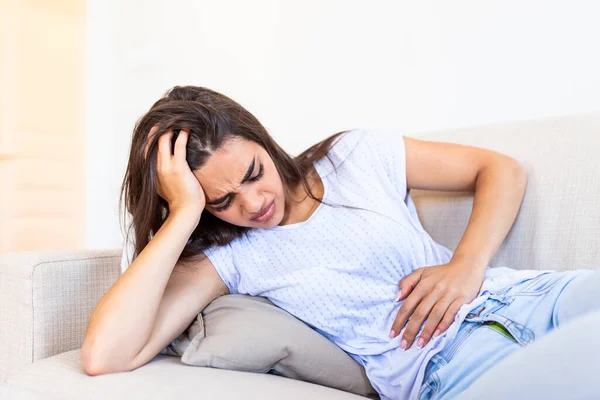 Woman in painful expression holding hands against belly suffering menstrual period pain, lying sad on home bed, having tummy cramp in female health concept