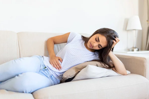 Woman in painful expression holding hands against belly suffering menstrual period pain, lying sad on home bed, having tummy cramp in female health concept