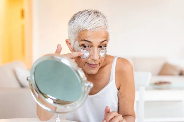 Under eye masks for puffiness, wrinkles, dark circles. Eye patches concept. Smiling senior gray haired woman in white shirt, applying silver eye patches while looking in the mirror