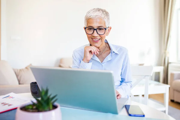 Focused old woman with white hair at home using laptop. Senior stylish entrepreneur wearing eyeglasses working on computer at home. Woman analyzing and managing domestic bills and home finance