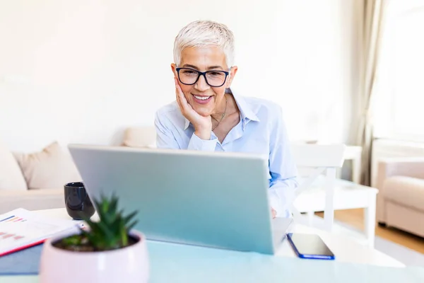 Focused old woman with white hair at home using laptop. Senior stylish entrepreneur wearing eyeglasses working on computer at home. Woman analyzing and managing domestic bills and home finance