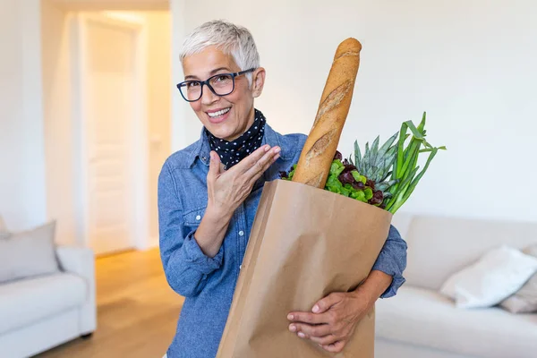 Happy mature woman holding a paper bag full of groceries from the supermarket. Elderly woman with groceries at home