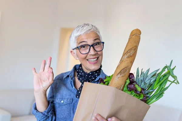 Elderly beautiful woman holding paper bag full of fresh healthy groceries and vegetables. showing OK sign