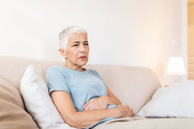 Senior woman suffering from stomach ache at home, unhappy older female sitting on bed, suffering from stomachache, food poisoning, gastritis, abdominal pain, climax