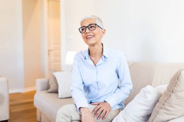 Older female with healthy smile sitting on couch at home. Smiling senior woman wearing glasses looking in distance.