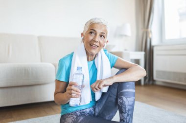 Elderly woman sitting on the mat, exhausted after the daily training. Senior woman taking a break while exercising at home. Athletic mature woman holding bottle of water