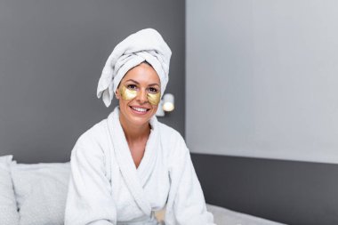 Beautiful smiling young woman in bathrobe and towel on head touching her face with Gold Cosmetics mask, Eye Patches. Spa, depilation and bodycare concept