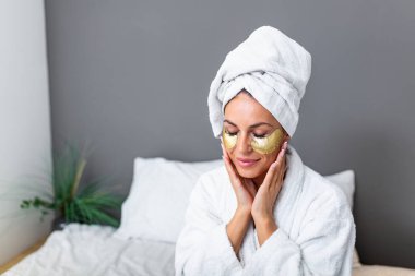 beautiful smiling young woman in bathrobe and towel on head touching her face with Gold Cosmetics mask, Eye Patches. Spa, depilation and bodycare concept