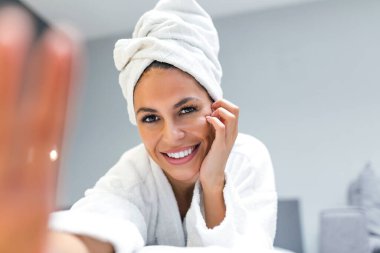 Smiling young woman lying on bed in bathrobe with her mobile phone taking a selfie.Beautiful woman relaxing on the bed after bath and looking at the phone camera taking a selfie