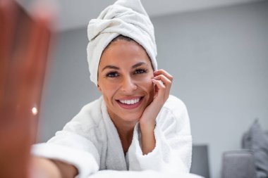 Smiling woman lying on bed in bathrobe with mobile phone taking a selfie. Beautiful woman relaxing on the bed after bath and looking at the phone camera taking a selfie