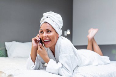 Young beautiful woman in towel and bathrobe sitting on bed at home and talking on her mobile smart phone. beautiful young woman wearing a white bathrobe, with a towel on her head in bed at home