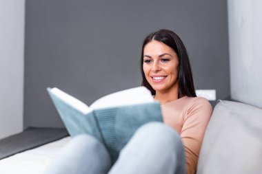 Genç ve güzel bir kadın kanepede uzanıyor ve evde kitap okuyor. Harika bir kitap. Güneşli bir sabahta kitap okuyan güzel genç bir kadın..